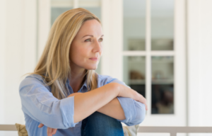 Kobieta siedząca w spokojnym, jasnym wnętrzu, patrząca w dal z wyrazem zadumy i nadziei.